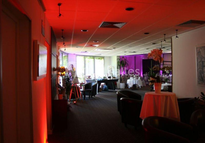 Salle lumineuse avec éclairage rouge et violet, tables rondes décorées de fleurs et fauteuils disposés autour.