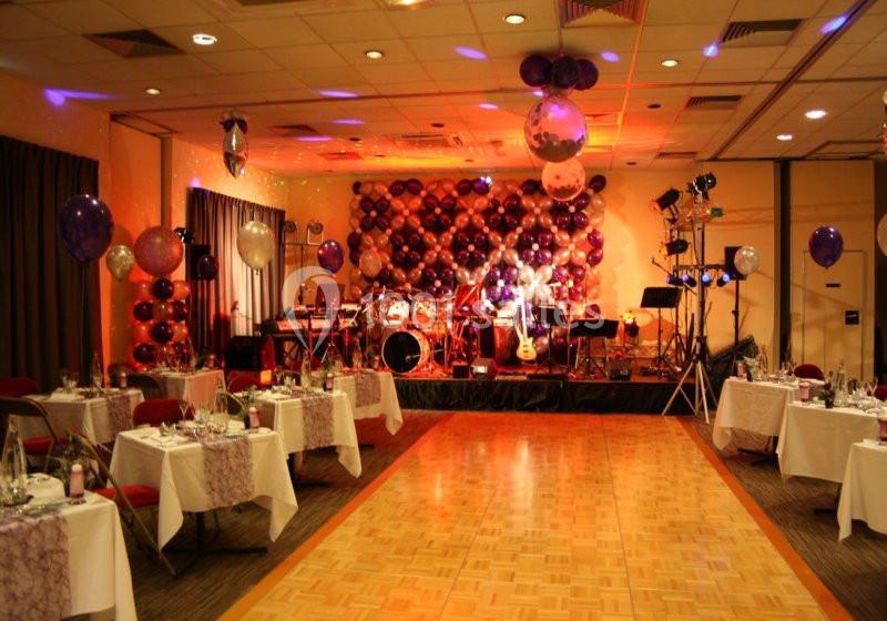 Salle décorée pour un événement avec tables dressées, piste de danse en bois et scène équipée d'instruments de musique.