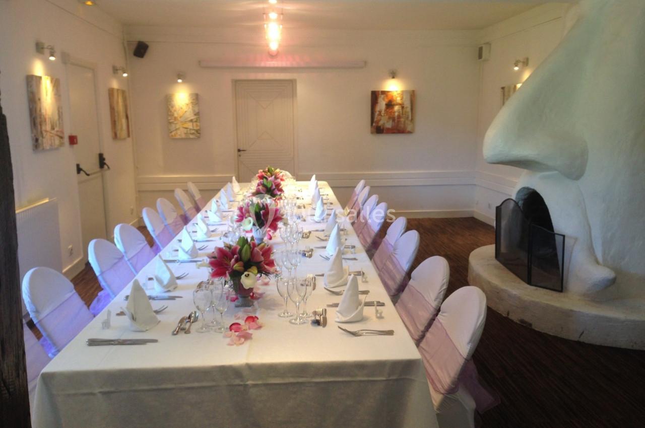 Table dressée pour un repas, décorée de fleurs, dans une salle lumineuse avec cheminée blanche et tableaux aux murs.