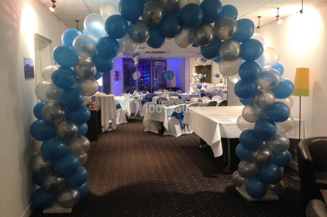 Arche décorative en ballons bleus, argentés et blancs dans une salle aménagée pour un événement festif.