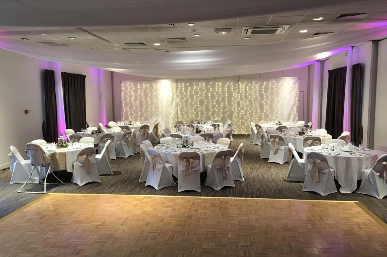 Salle de réception décorée avec des tables rondes dressées, des chaises couvertes et un fond lumineux avec guirlandes.