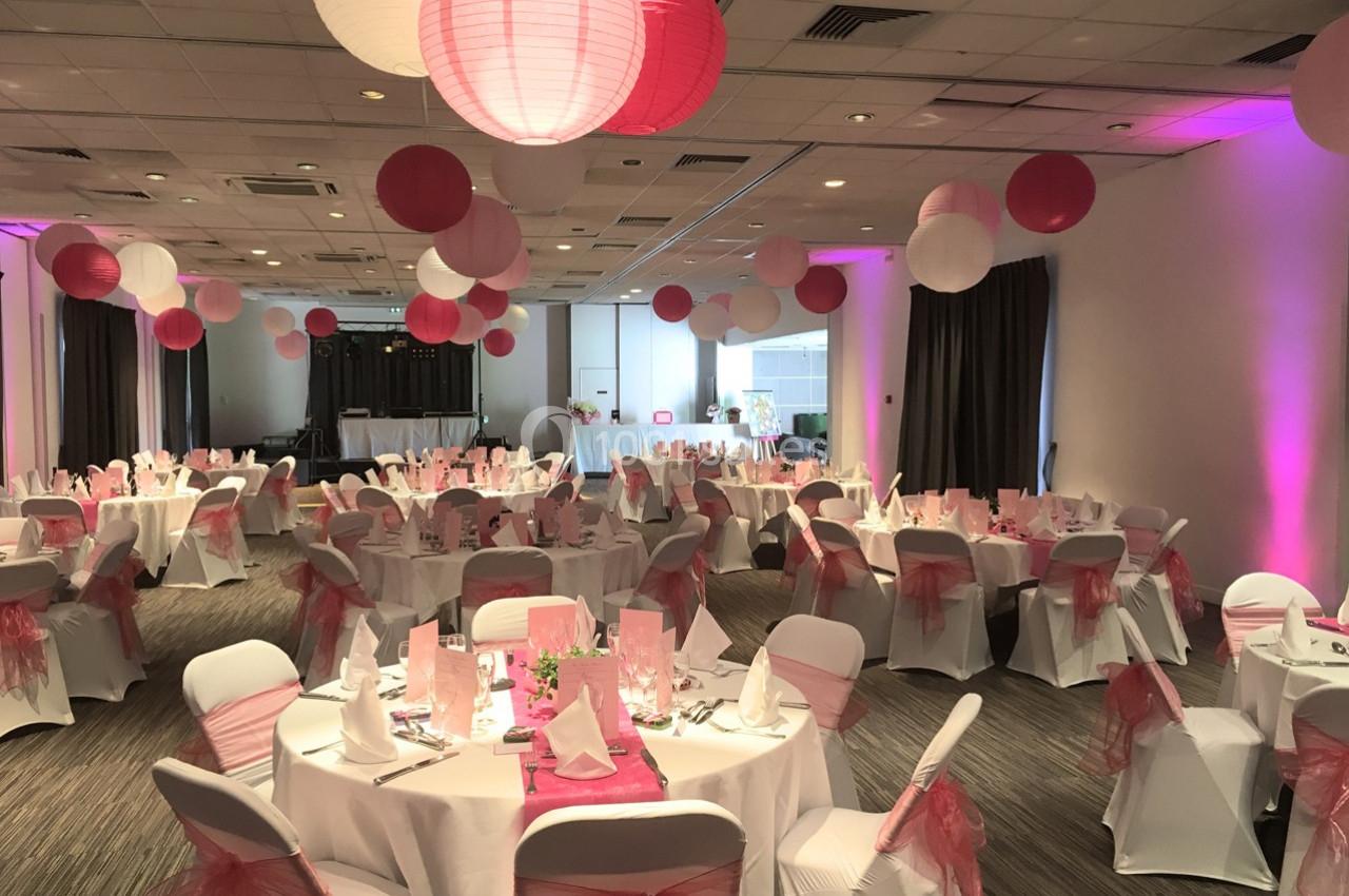 Salle de réception décorée avec des lanternes roses et blanches, tables rondes dressées et chaises ornées de nœuds roses.
