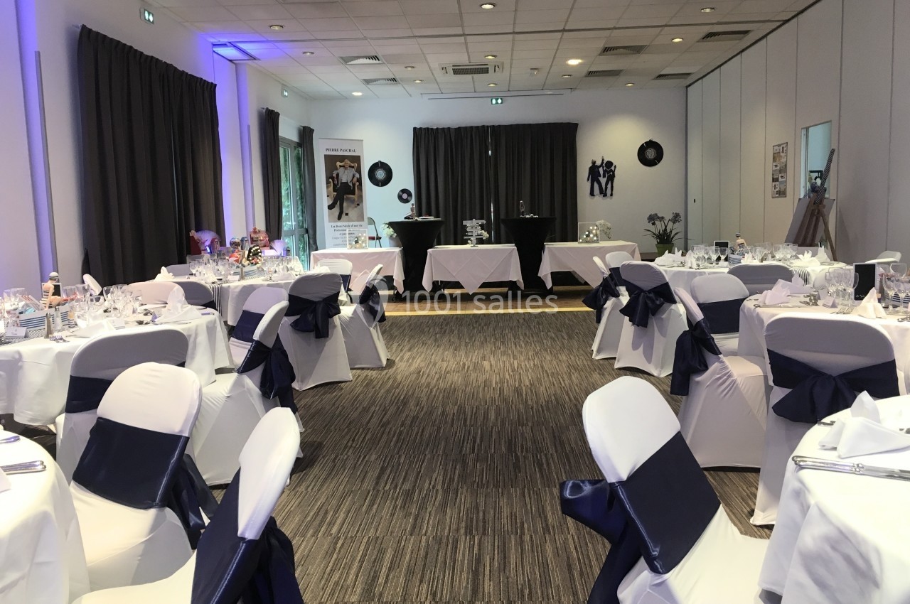 Salle de réception décorée avec des tables dressées, nappes blanches et nœuds bleu marine sur les chaises.