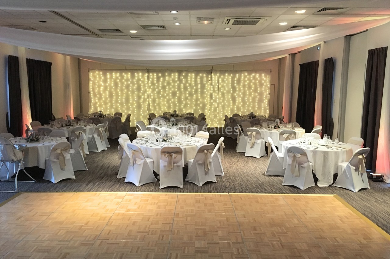 Salle de réception décorée avec des tables rondes dressées et un mur illuminé de guirlandes lumineuses.
