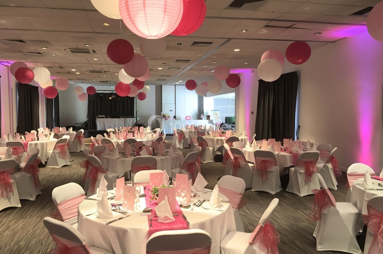 Salle de réception décorée avec des lanternes roses et blanches, tables rondes dressées et chaises ornées de nœuds roses.