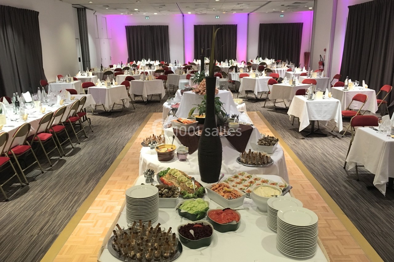 Salle de réception avec tables dressées et buffet central composé de plats variés, entouré de chaises rouges.