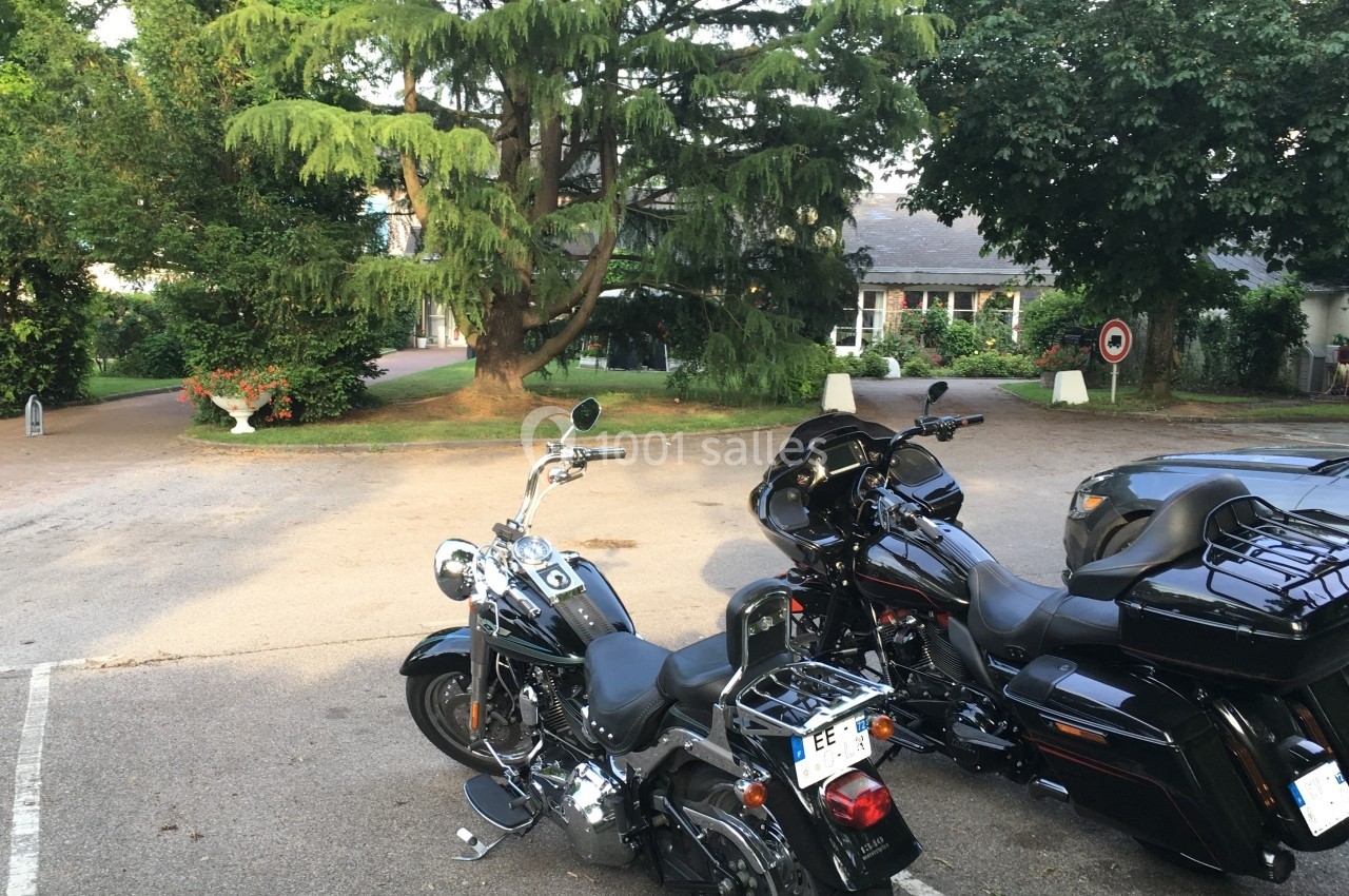Deux motos stationnées sur un parking entouré d'arbres et de végétation, avec un bâtiment en arrière-plan.