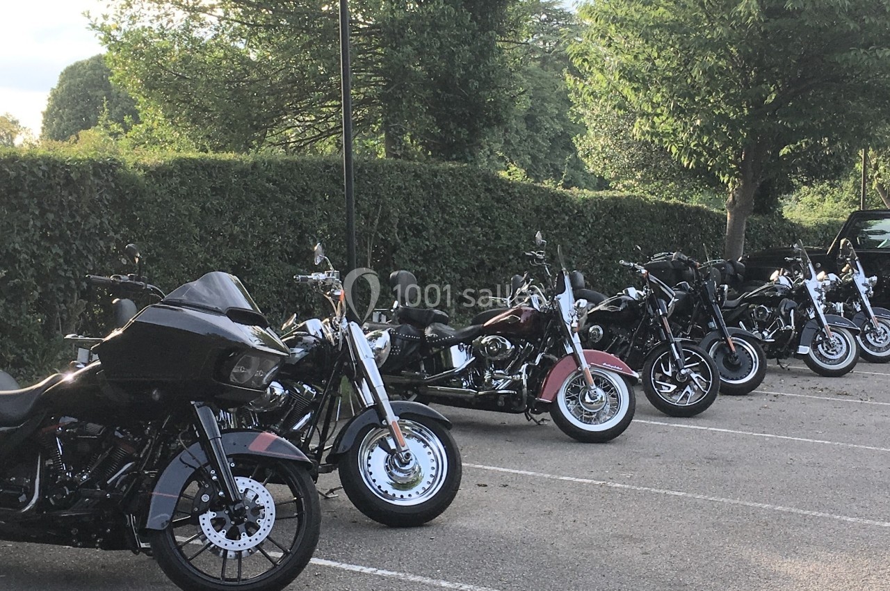 Plusieurs motos garées en ligne dans un parking extérieur bordé de haies et d'arbres.