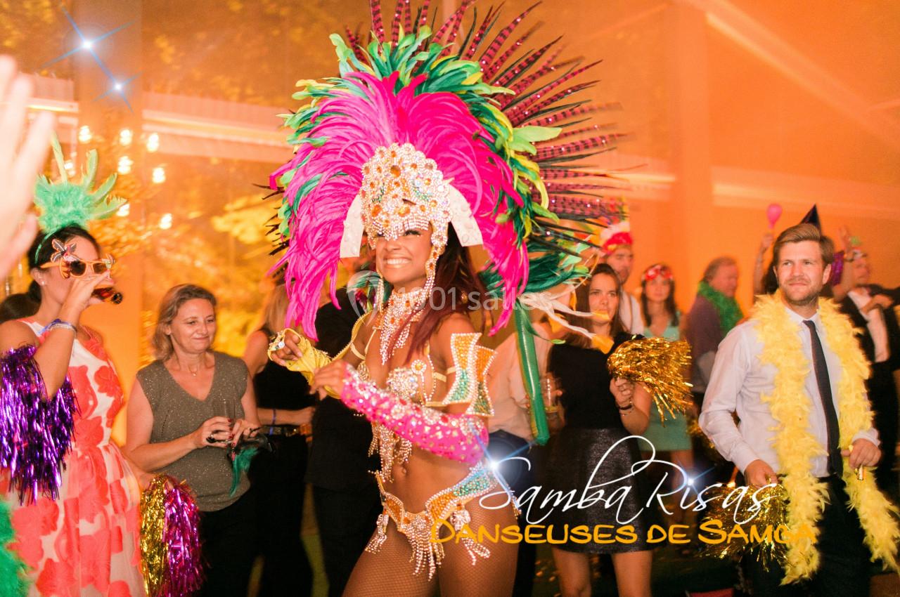 Danseuse de samba en costume coloré avec plumes, entourée de spectateurs lors d'une fête festive en intérieur.