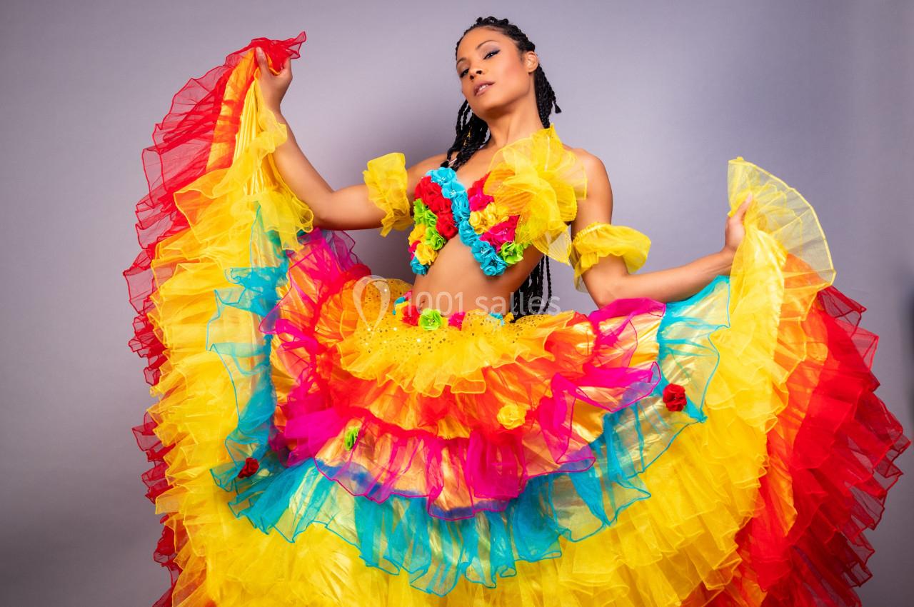 Femme portant une robe colorée à volants rouges, jaunes et bleus, posant devant un fond gris.