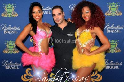 Trois hommes souriants en tenue de capoeira avec des drapeaux brésiliens, posant dans un parc ensoleillé.