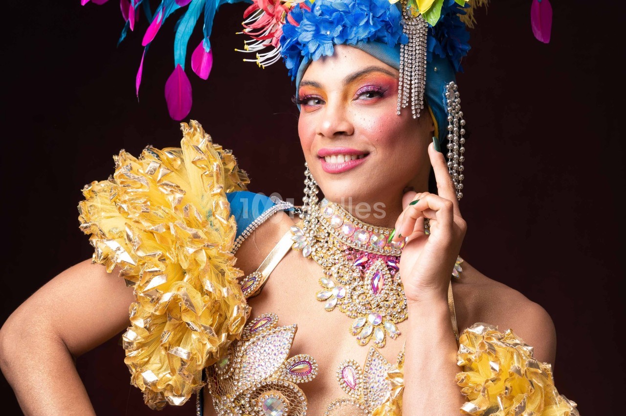 Personne souriante en costume coloré avec plumes, perles et ornements, posant sur un fond sombre.