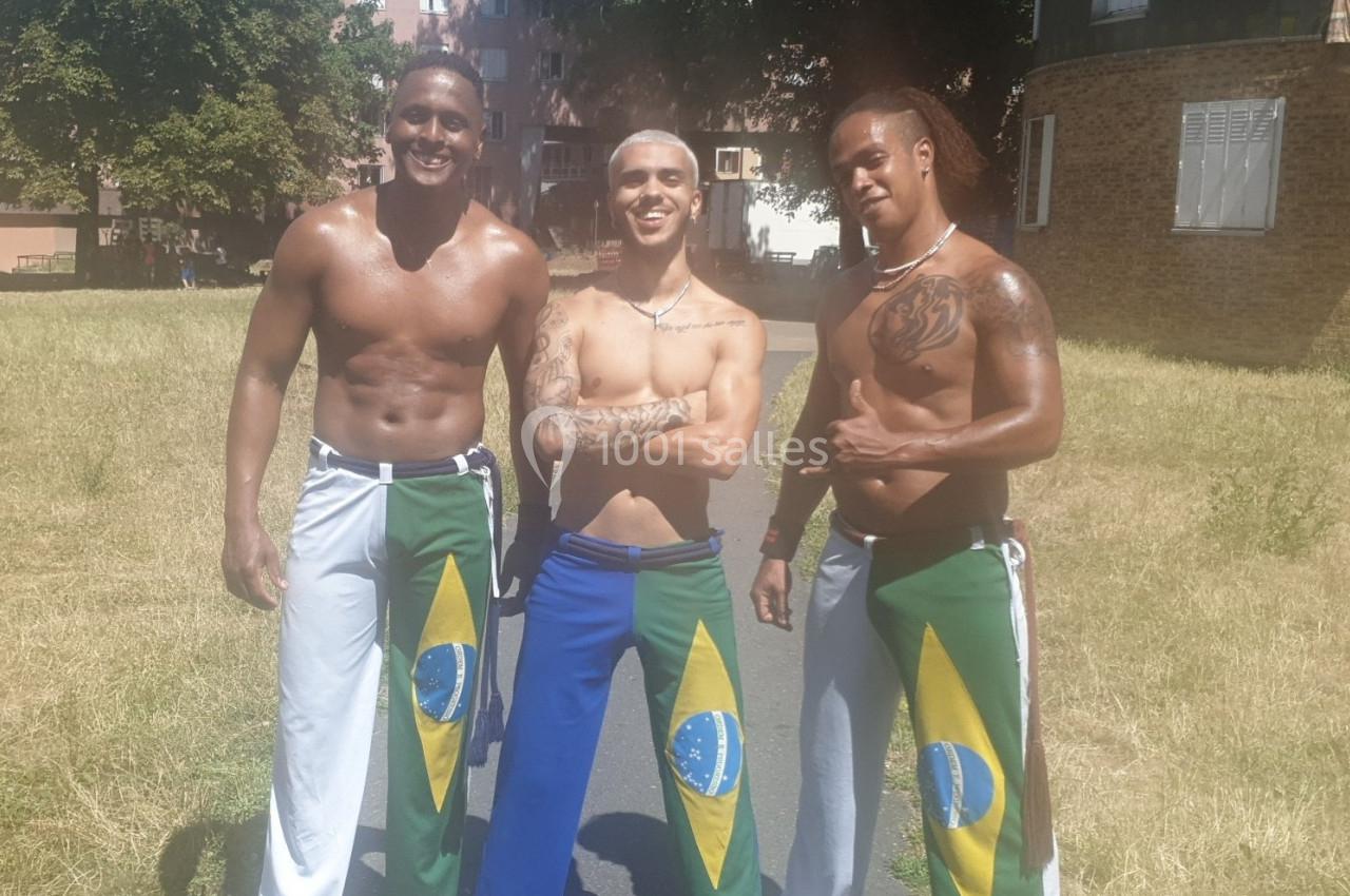 Trois hommes souriants en tenue de capoeira avec des drapeaux brésiliens, posant dans un parc ensoleillé.