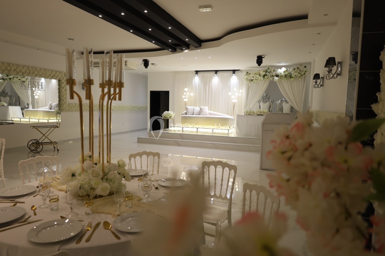 Salle de réception élégante avec tables décorées de fleurs blanches et un espace scénique illuminé en arrière-plan.