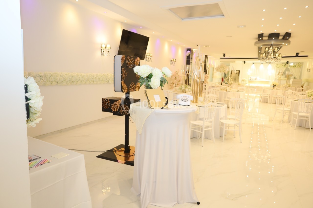 Salle de réception lumineuse avec tables décorées, fleurs blanches et un coin photo équipé d'accessoires.