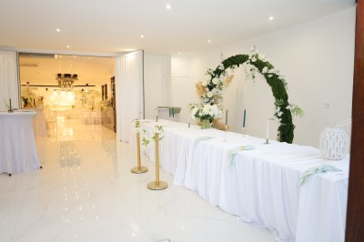 Salle lumineuse avec table drapée de blanc, décorée de fleurs et bougies, donnant sur un extérieur arboré.