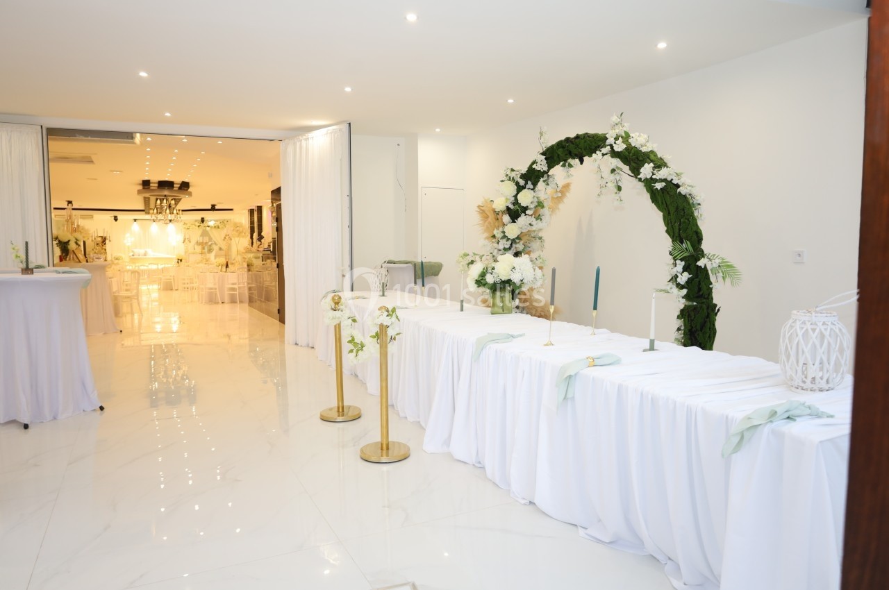 Salle de réception lumineuse avec tables décorées de nappes blanches et arche florale ornée de verdure et fleurs blanches.