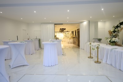 Salle lumineuse avec table drapée de blanc, décorée de fleurs et bougies, donnant sur un extérieur arboré.