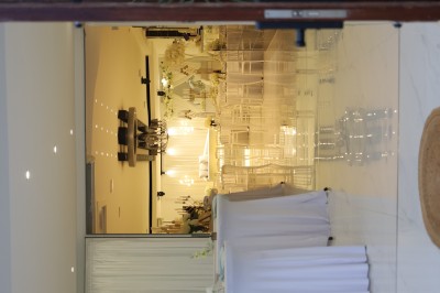 Salle lumineuse avec table drapée de blanc, décorée de fleurs et bougies, donnant sur un extérieur arboré.