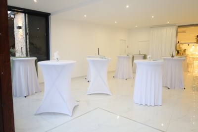 Salle lumineuse avec table drapée de blanc, décorée de fleurs et bougies, donnant sur un extérieur arboré.