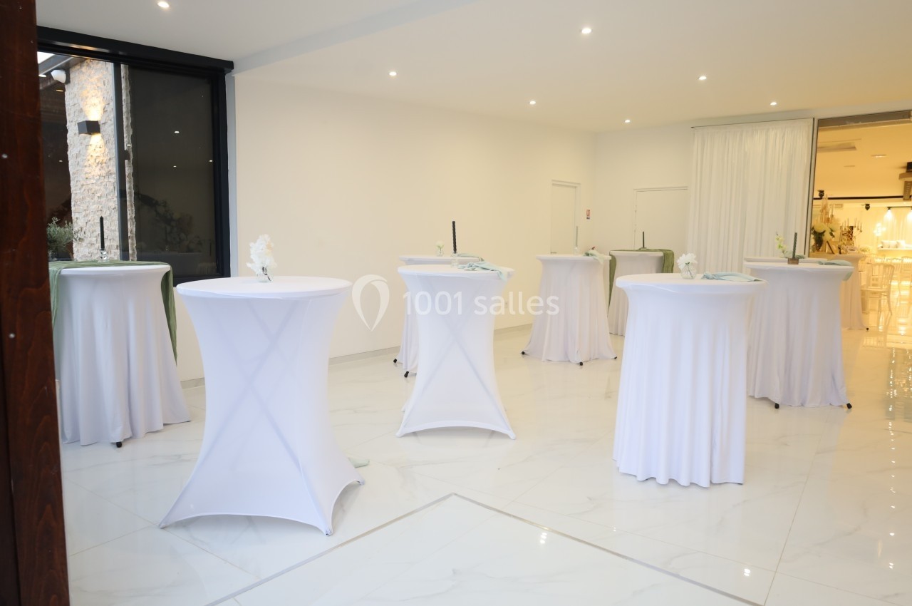 Salle lumineuse avec tables hautes recouvertes de nappes blanches, disposées pour un événement ou une réception.
