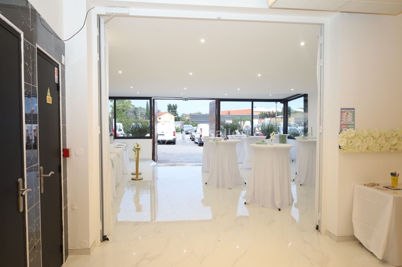 Salle lumineuse avec tables hautes dressées, sol en marbre blanc et vue sur un parking à travers de grandes baies vitrées.