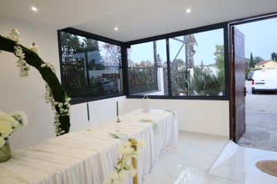 Salle lumineuse avec table drapée de blanc, décorée de fleurs et bougies, donnant sur un extérieur arboré.