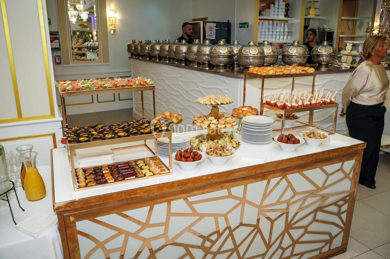 Buffet varié avec pâtisseries, amuse-bouches, pains et boissons, présenté sur une table décorée dans un cadre élégant.