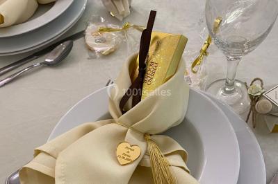 Table dressée avec une assiette blanche, un cadeau doré, une serviette beige nouée et des macarons en décoration.