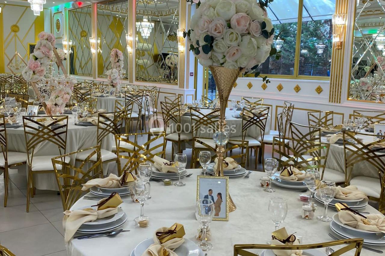 Salle de réception élégante avec tables décorées de nappes blanches, chaises dorées et centres de table floraux.