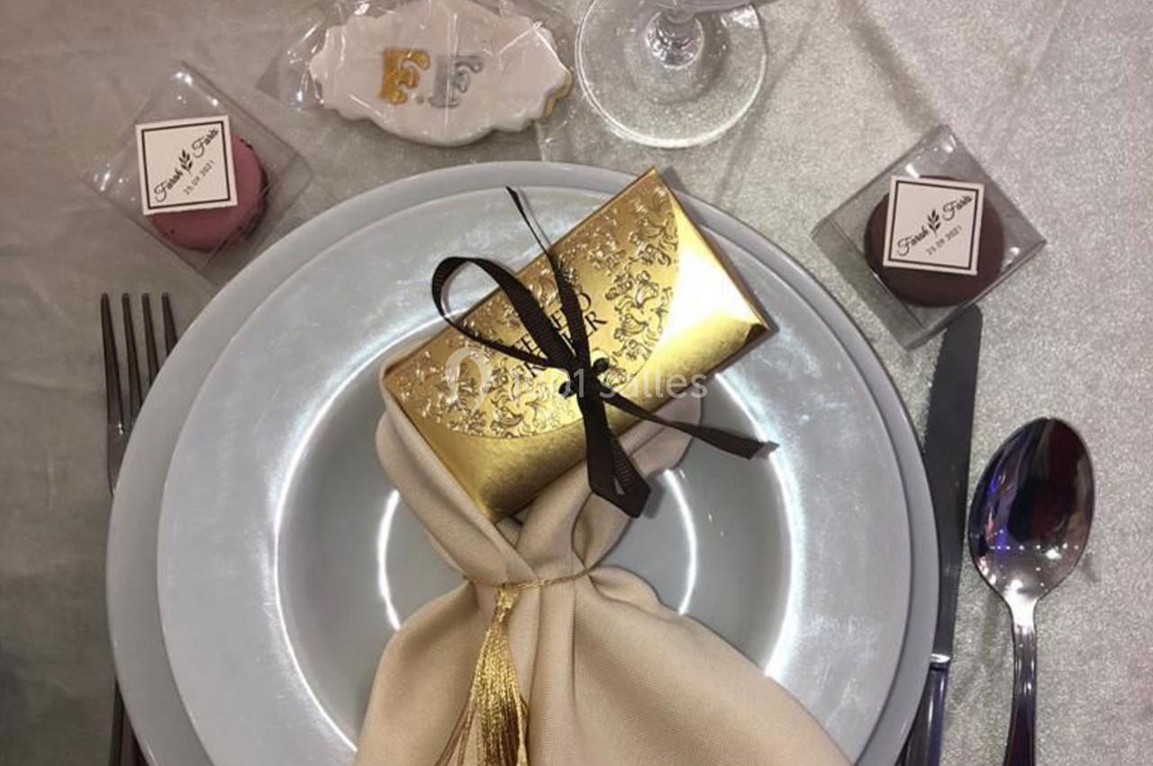 Table dressée avec une assiette blanche, un cadeau doré, une serviette beige nouée et des macarons en décoration.