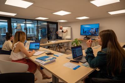 Salle de classe vide avec des tables et chaises jaunes alignées, éclairée par des fenêtres sur les côtés.