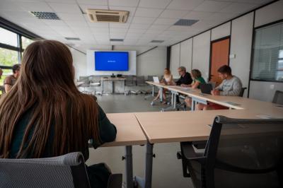 Salle de classe vide avec des tables et chaises jaunes alignées, éclairée par des fenêtres sur les côtés.