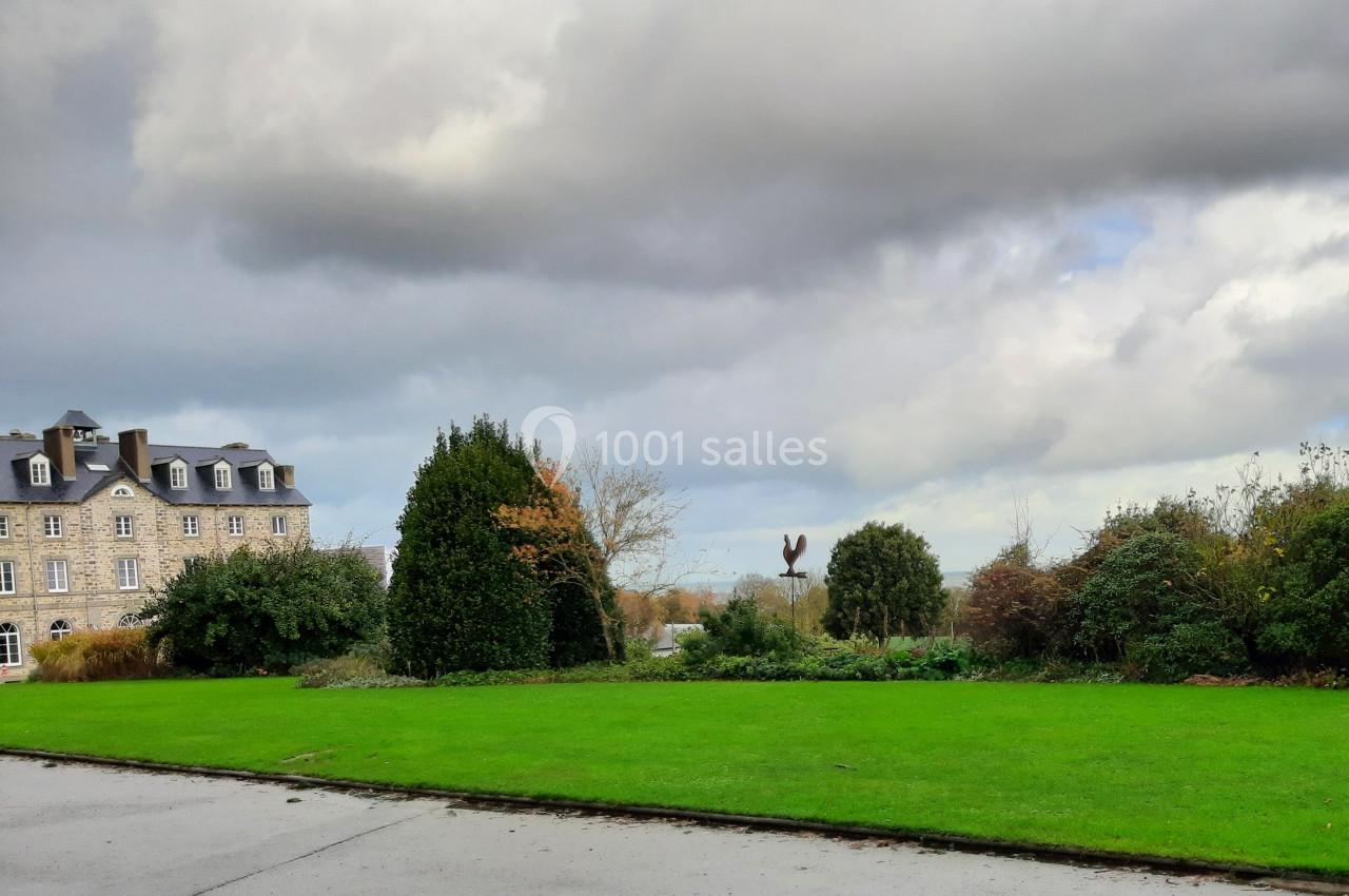 Pelouse verte bordée d'arbustes et d'arbres, avec un bâtiment en pierre à gauche et un ciel nuageux en arrière-plan.