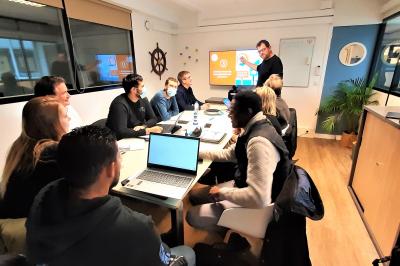 Salle de classe vide avec des tables et chaises jaunes alignées, éclairée par des fenêtres sur les côtés.