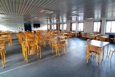 Salle de classe vide avec des tables et chaises jaunes alignées, éclairée par des fenêtres sur les côtés.
