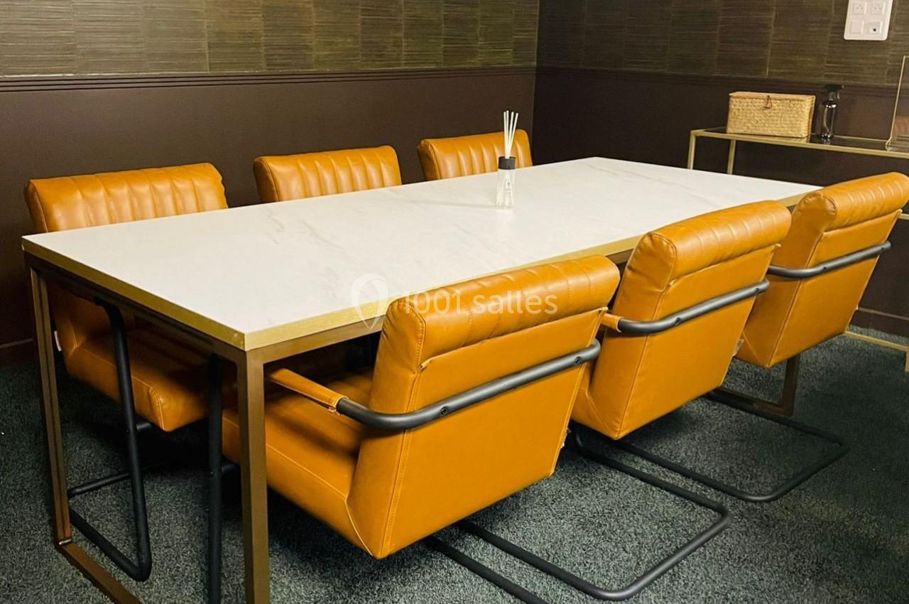 Salle de réunion avec table blanche, six chaises en cuir orange et mur en bois sombre.
