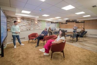 Salle moderne avec tables noires, chaises bleues, étagères murales, et écran mural dans un espace lumineux et décoré.