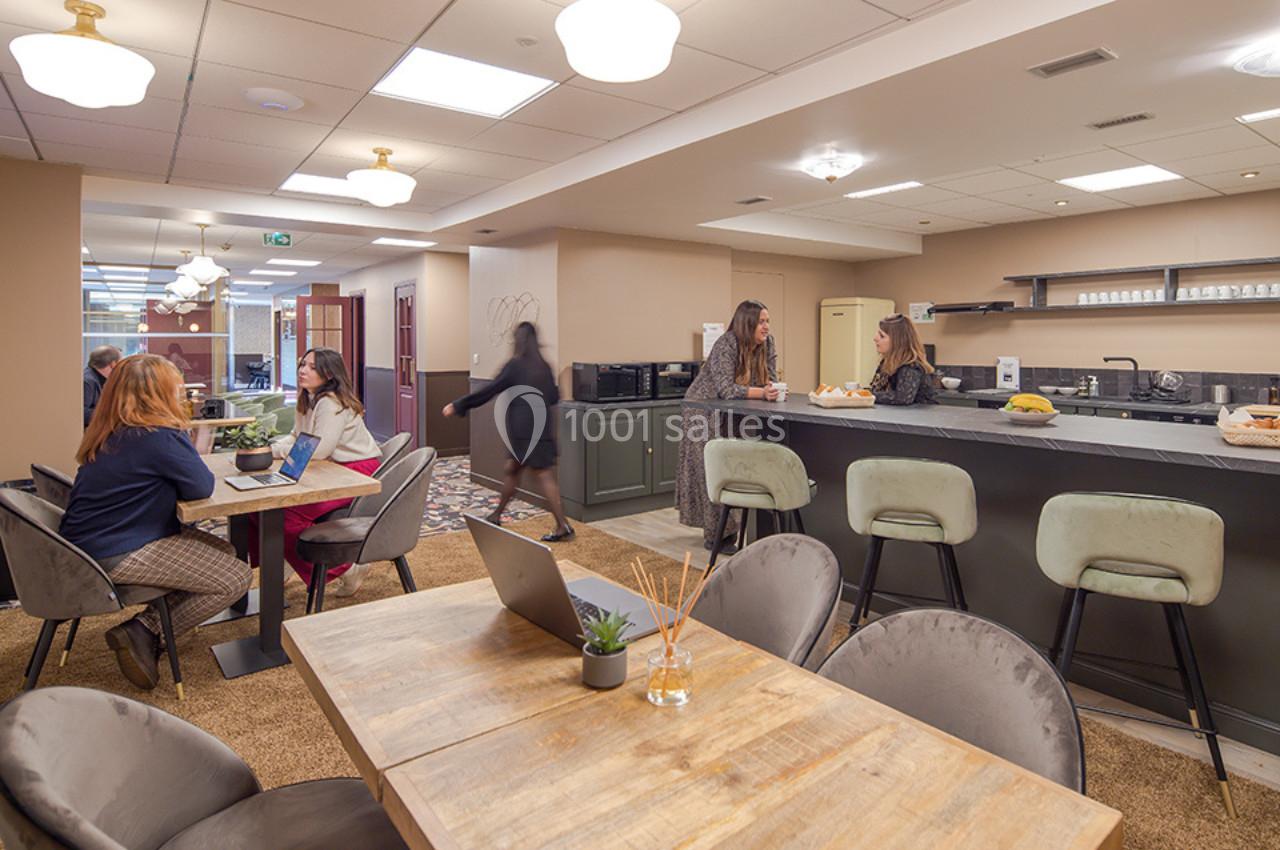 Espace de coworking avec tables, chaises, cuisine équipée et plusieurs personnes discutant ou travaillant.