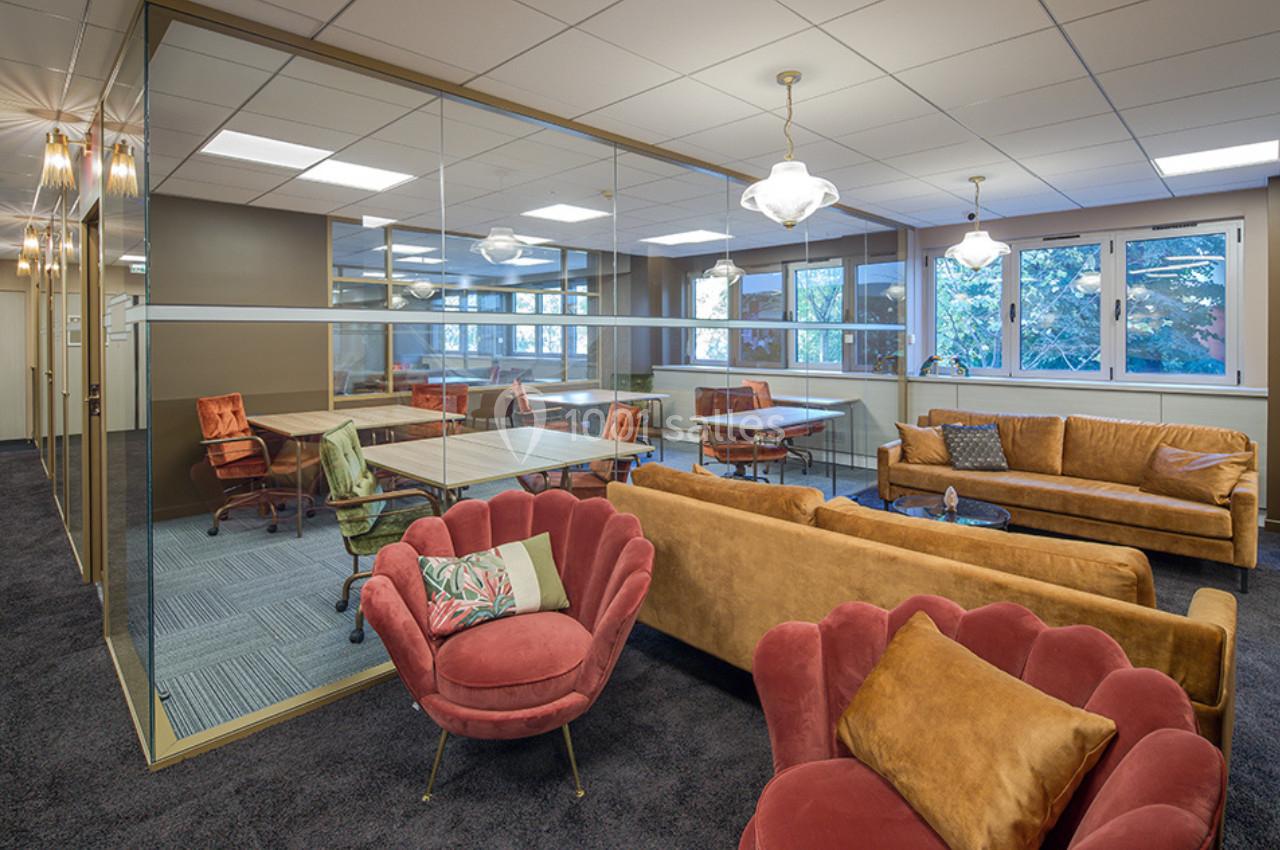 Espace de coworking lumineux avec mobilier coloré, canapés, fauteuils et cloison vitrée séparant une salle de réunion.