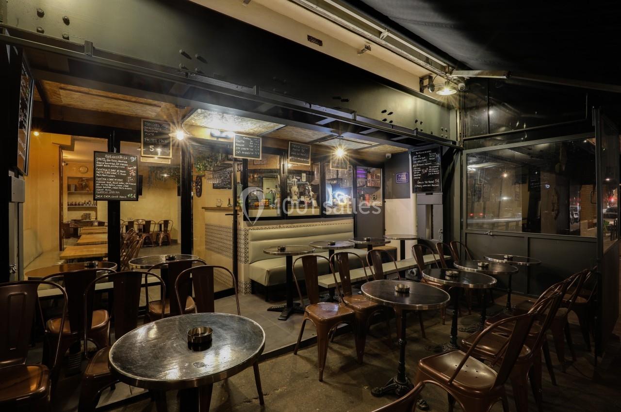Terrasse de café vide en soirée, avec des tables en métal et un intérieur éclairé visible à travers une ouverture.