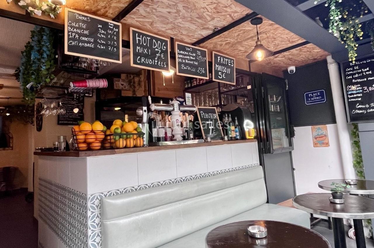Comptoir de bar avec fruits frais, ardoises de menus suspendues et tables métalliques dans un espace convivial.