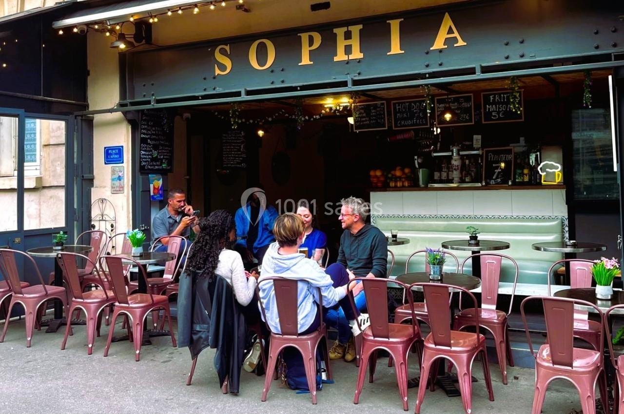 Terrasse d'un café avec des clients assis autour de tables, devant une façade portant l'enseigne ’Sophia’.