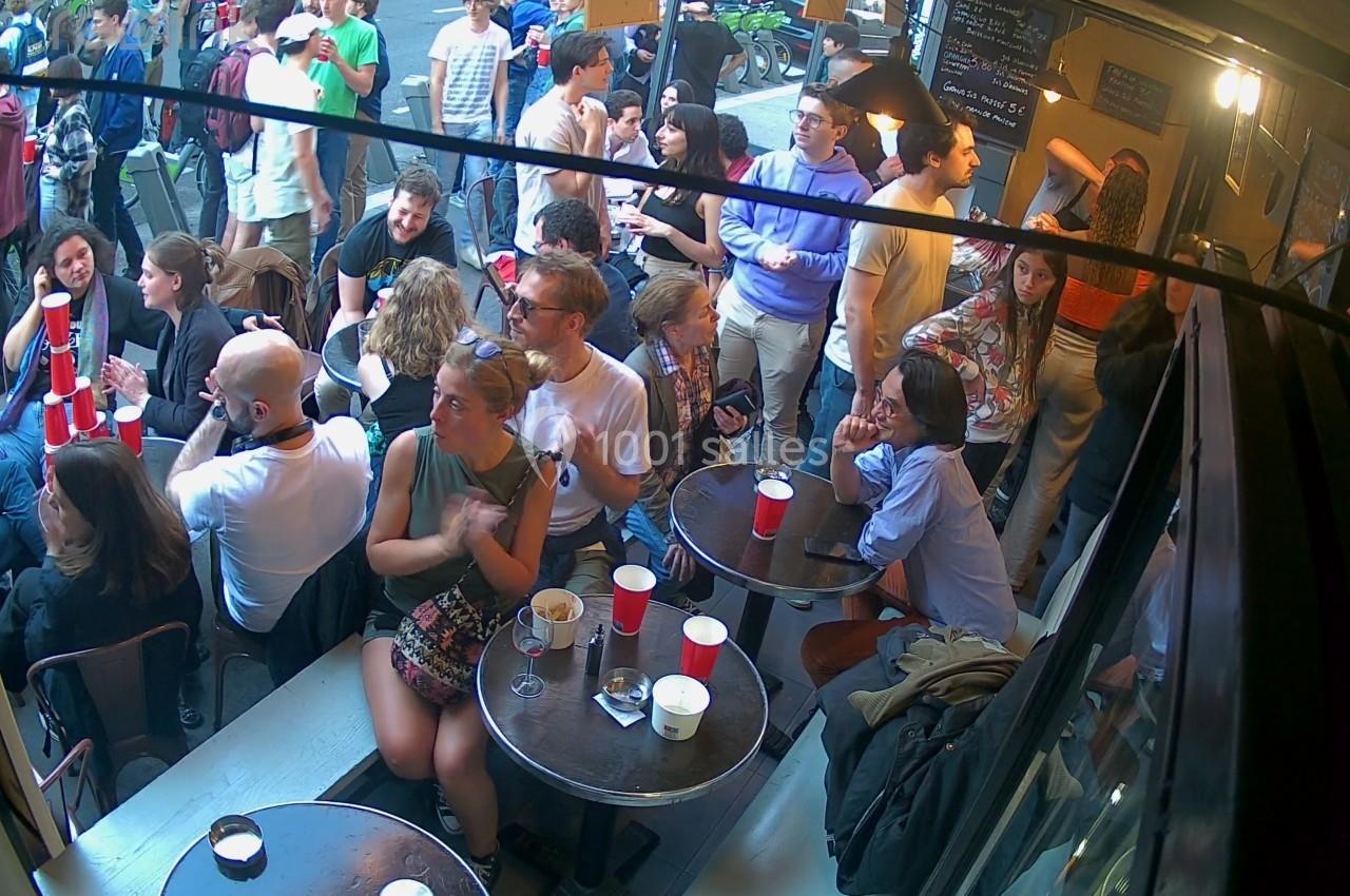 Groupe de personnes assises et debout dans un café animé, avec des boissons sur les tables et une ambiance conviviale.