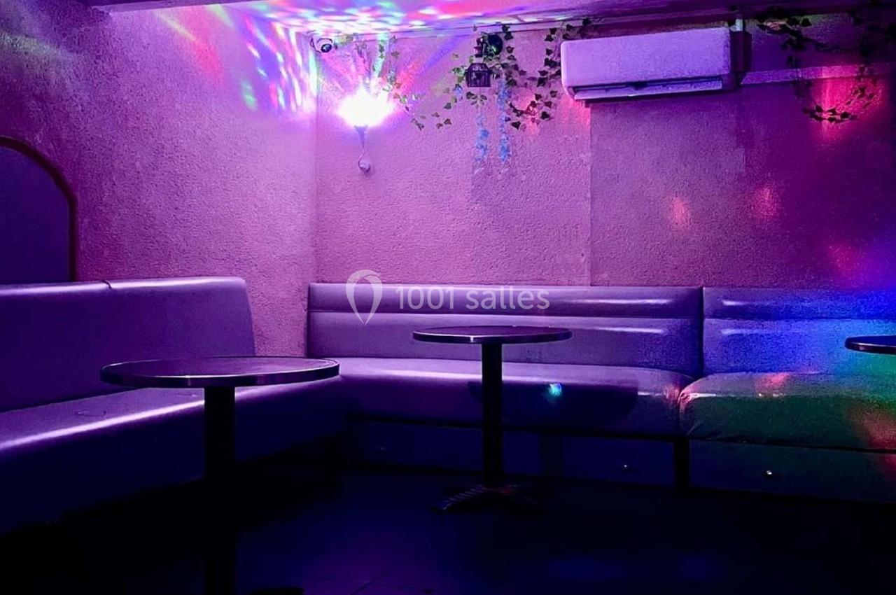 Salle avec banquettes violettes, tables rondes noires et éclairage coloré diffus sous une lampe suspendue.