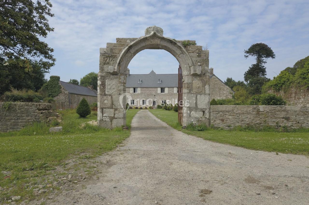 Portail en pierre ancienne encadrant une allée menant à une maison en pierre entourée de verdure.