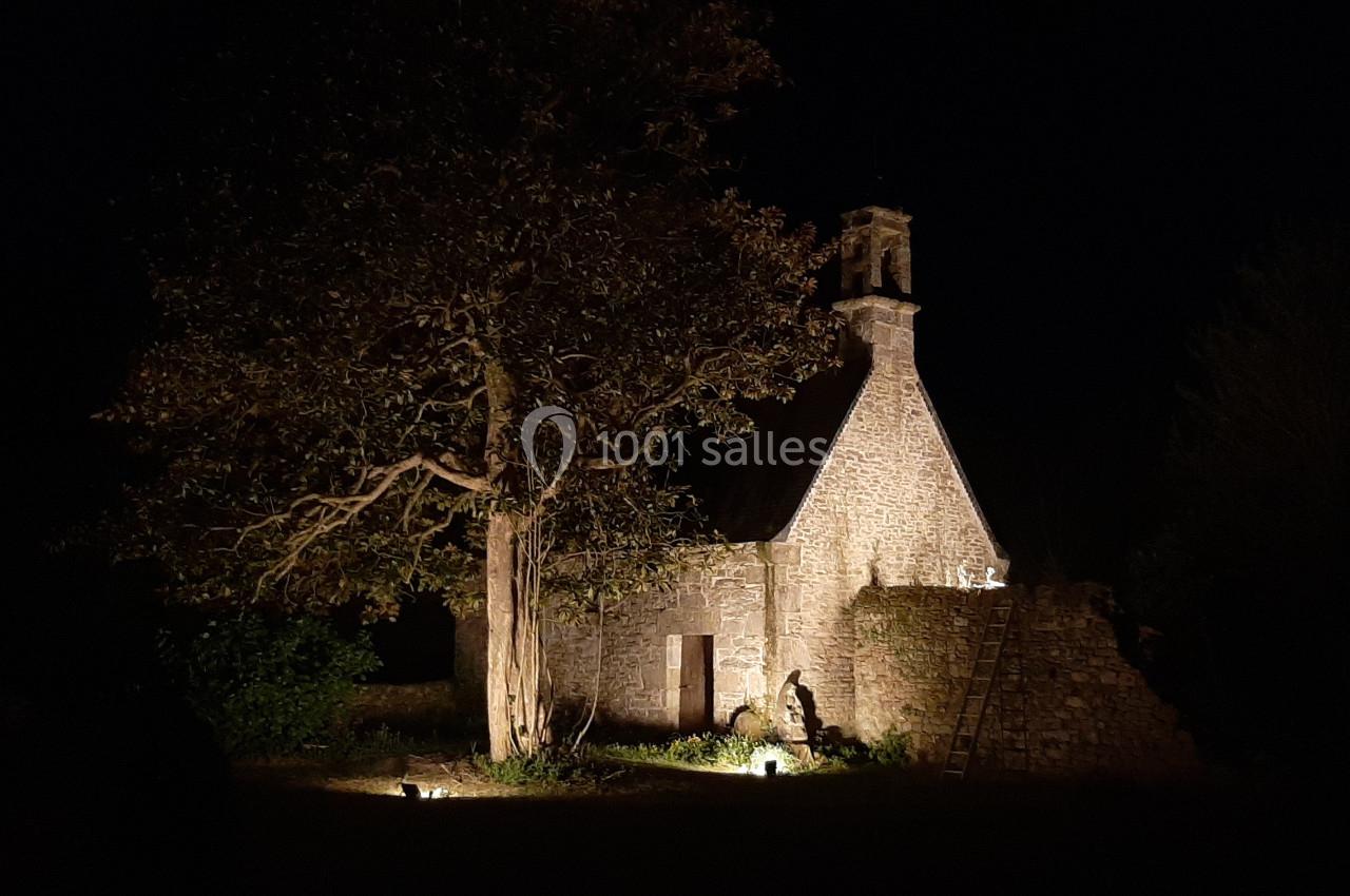 Église en pierre éclairée de nuit, entourée d'arbres et d'un jardin sombre.