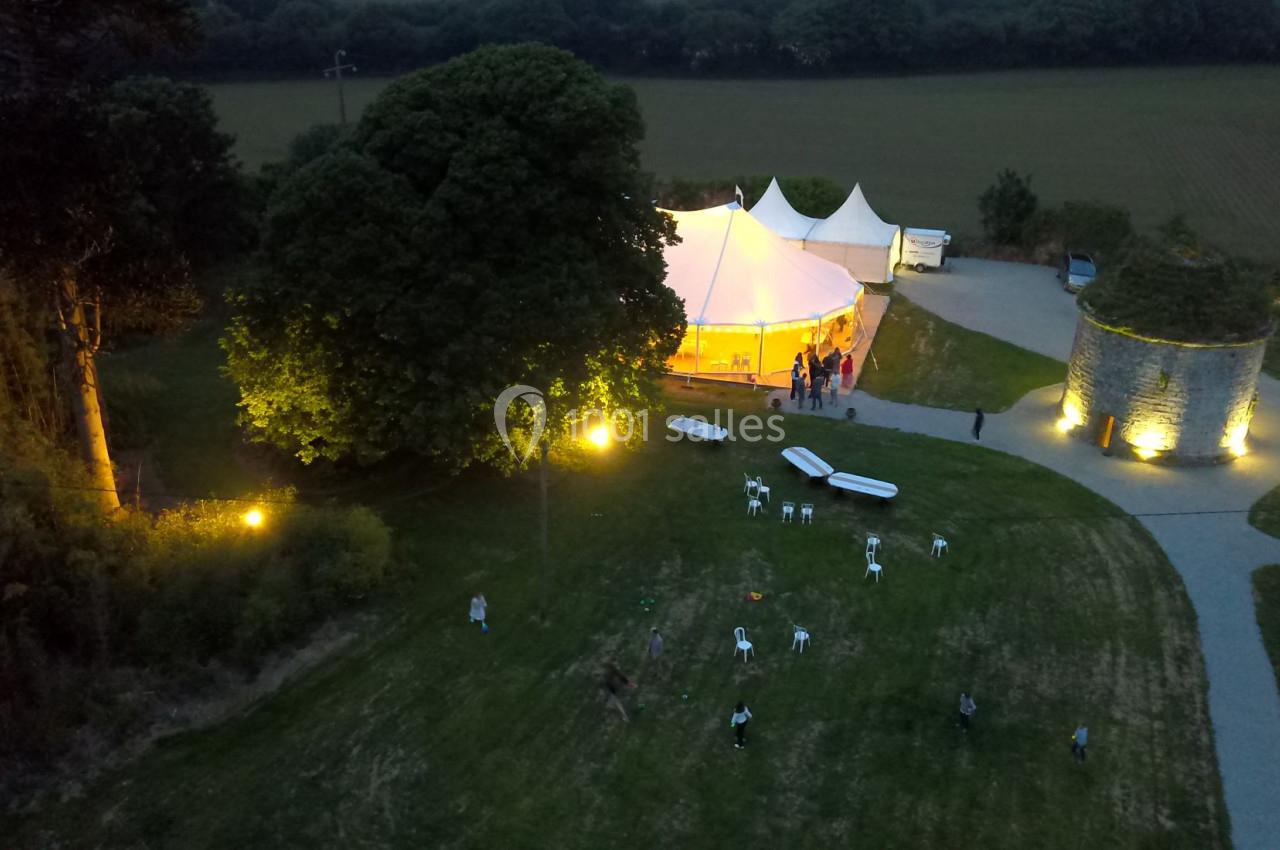 Vue aérienne d'une tente éclairée pour un événement nocturne, entourée de pelouses, d'arbres et de structures en pierre.
