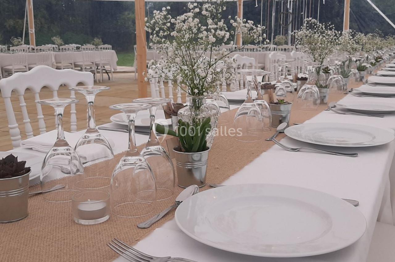 Tables de réception alignées avec nappes blanches, vaisselle, verres, et décorations florales dans un cadre extérieur.