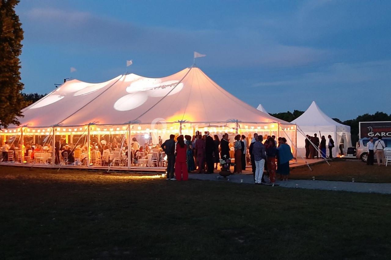 Grande tente éclairée en soirée, accueillant des invités debout et assis dans un cadre extérieur verdoyant.